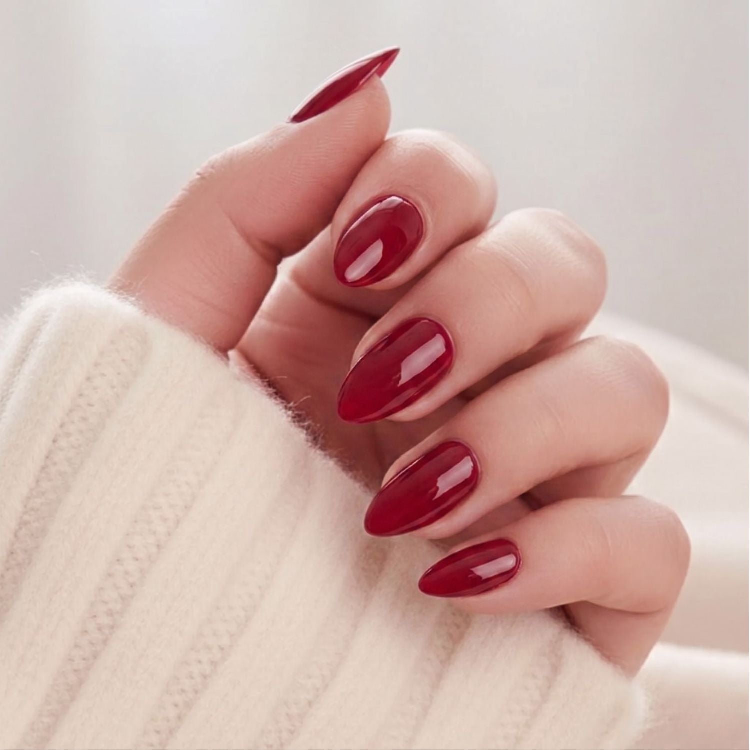 Ruby Romance press-on nagels in glanzend donkerrood, amandelvormig. Op een hand in zachte gebreide trui