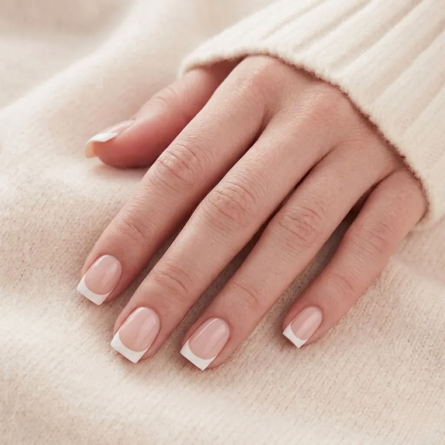 Angelic Aura press-on nagels op een hand tegen gebreide trui. Korte vierkante Franse manicure met nude basis en witte tip