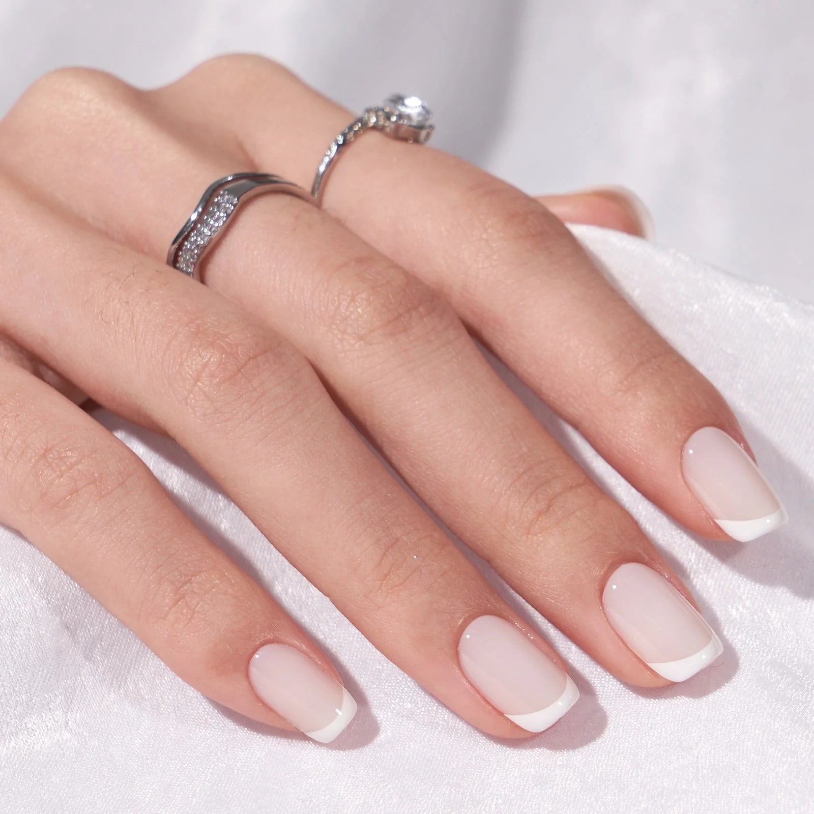 Frosted French gelnagels close-up – vierkante Franse manicure met zilveren ringen tegen witte stof