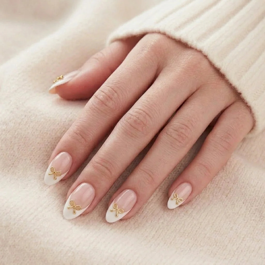 Blushing Bow nagels op hand – amandelvormige press-on nagels met franse witte tip en 3D gouden strik tegen beige gebreide trui