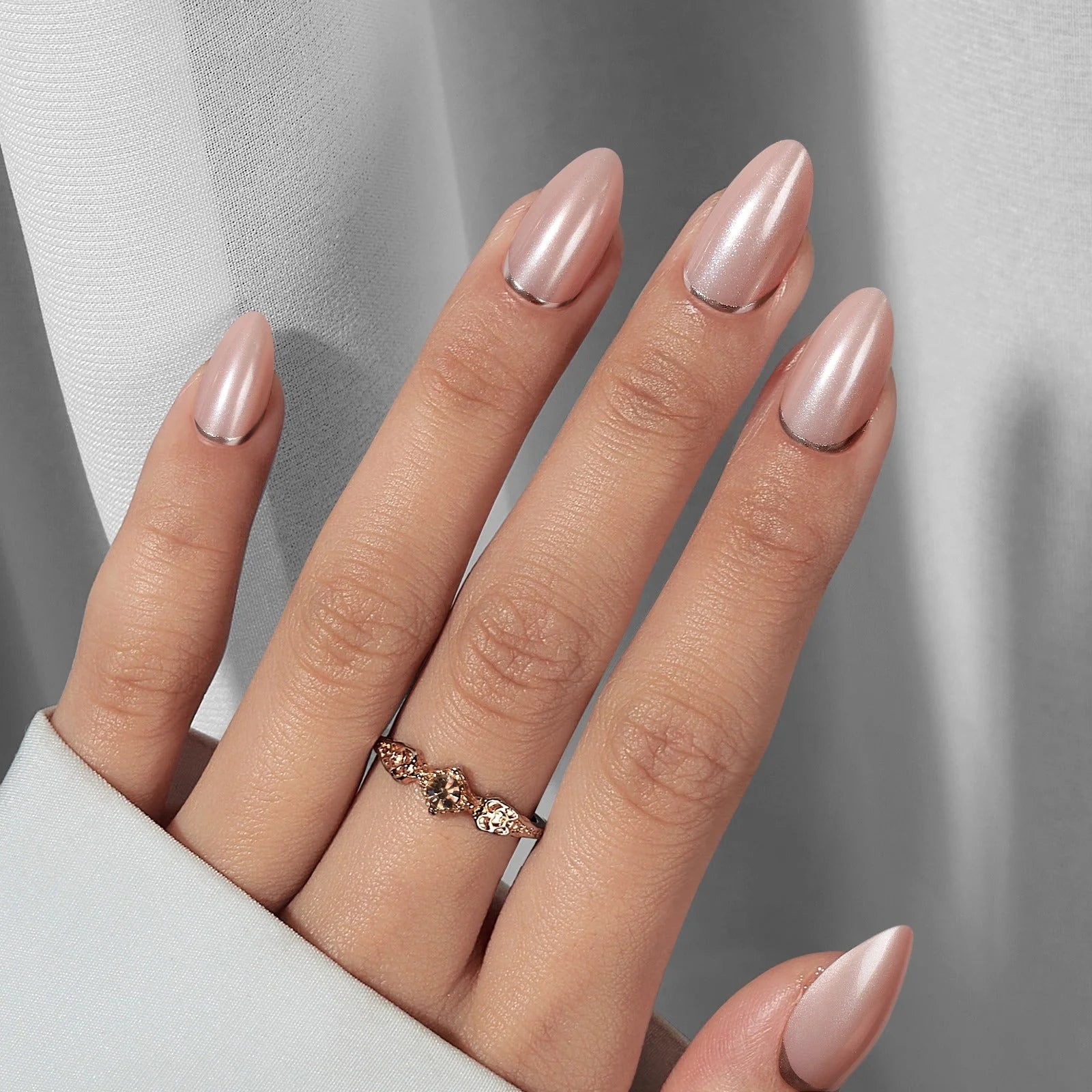 Amandelvormige plaknagels met parelglans – Shimmering Silver met roségouden ring tegen grijs zijden gordijn