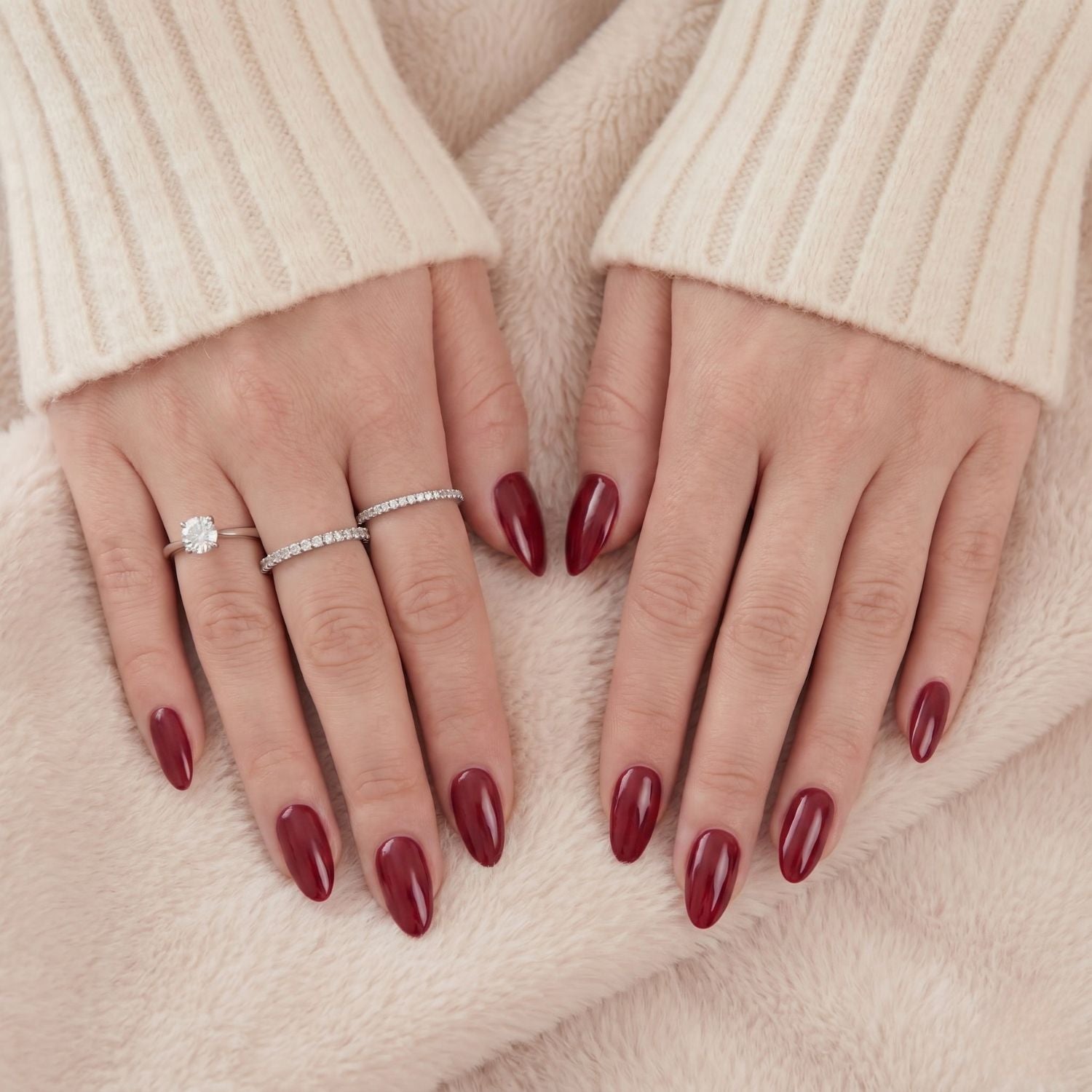 Ruby Romance donkerrode press-on nagels in amandelvorm. Handen van model met zilveren ringen op een zachte lichte deken