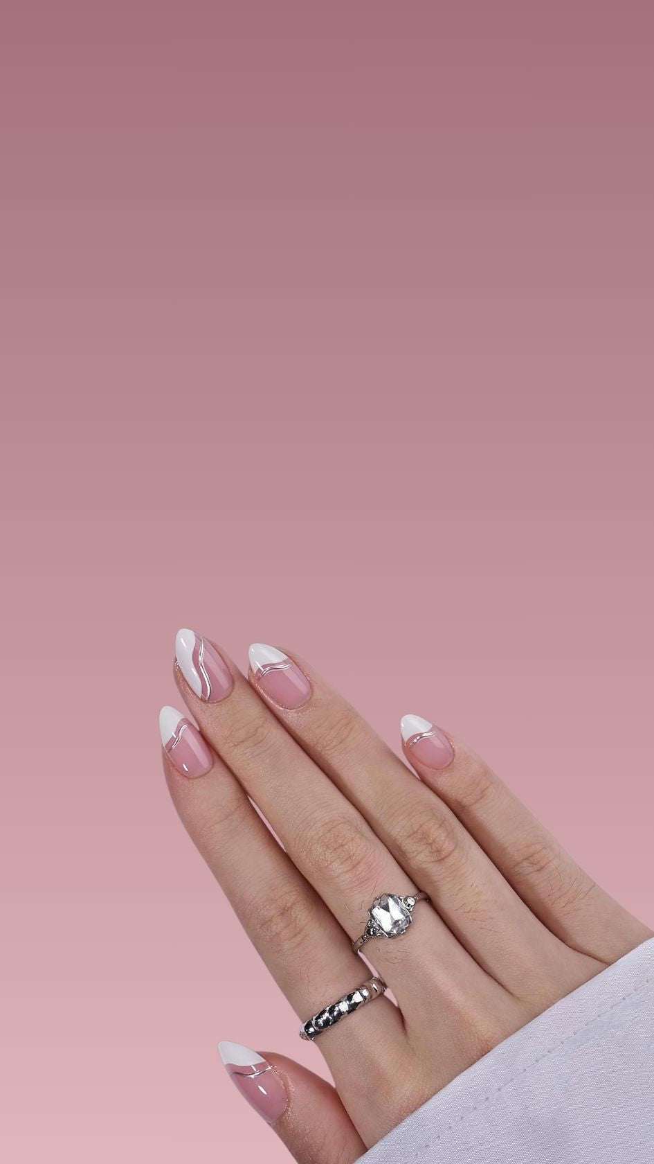 Hand met amandelvormige press-on nagels, nudroze basis en witte tips met elegante golvende zilveren lijn. Twee zilveren ringen
