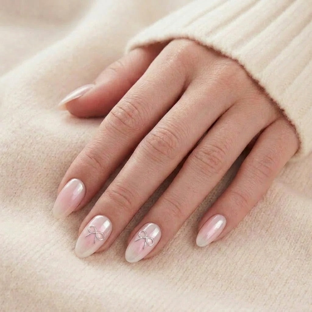 Aura Amoré press-on nagels in parelmoer-chrome met roze blush-ombré en zilveren strik op amandelnagels tegen een beige gebreide trui