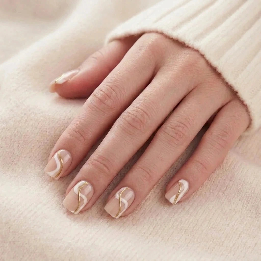 Korte, beige nepnagels met wit en goudkleurig golfdesign op een modelhand tegen een gebreide trui.