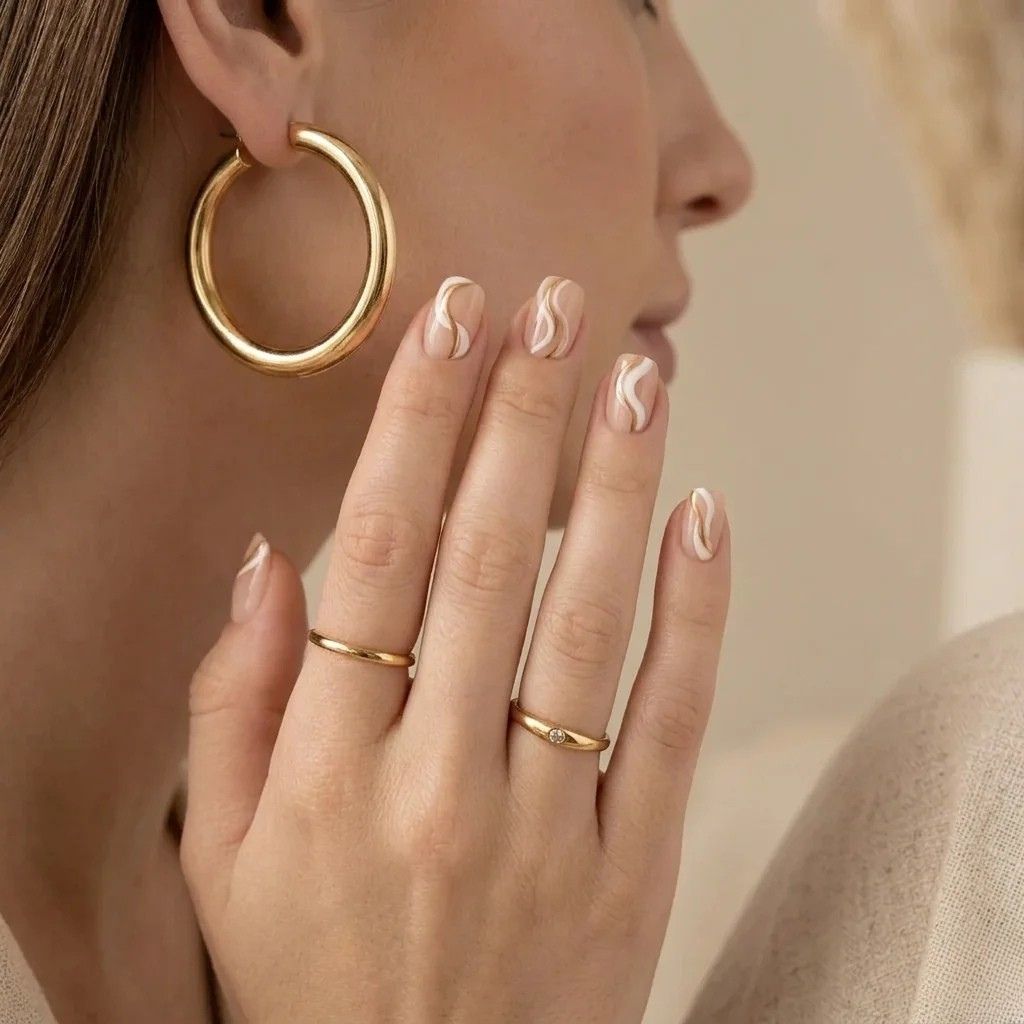 Korte, afgeronde press-on nagels in nude met witte en beige golvende lijnen. Hand met gouden ringen.