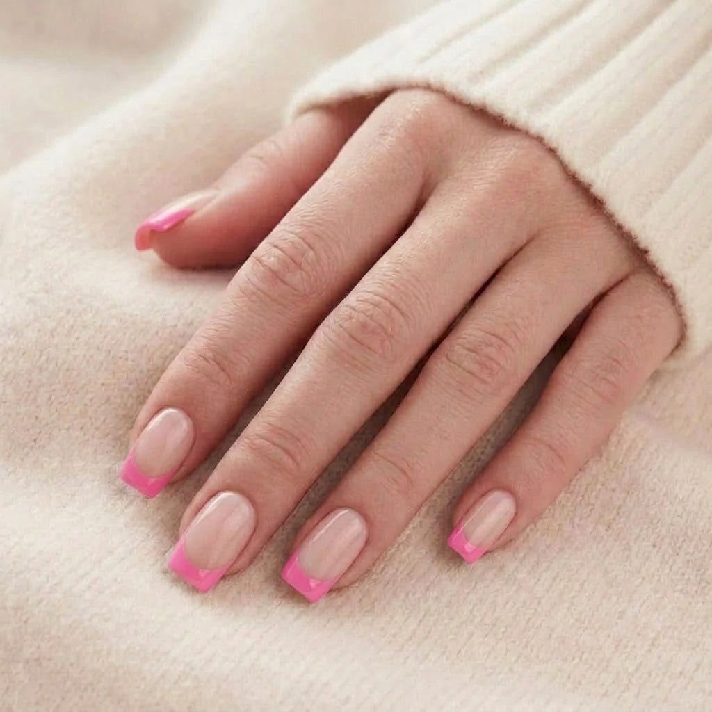 Pixie Pink press-on nagels in kort vierkant met nude basis en helderroze French tip op een hand tegen een beige gebreide trui